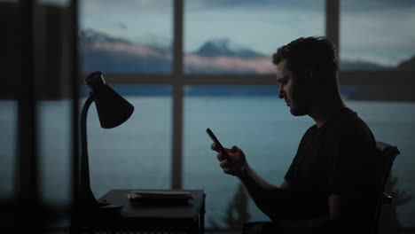 The-silhouette-of-a-thoughtful-man-sitting-at-a-table-against-the-background-of-a-window-with-oceans-and-the-sea.-A-restless-man-sitting-at-a-table-with-a-desk-lamp